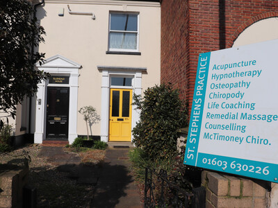 Cream‑coloured building with a bright yellow door, in front a small front garden with paving stones, gravel, shrubs, and a narrow path. In the foreground, a large turquoise and white sign reads ‘St. Stephen’s Practice’